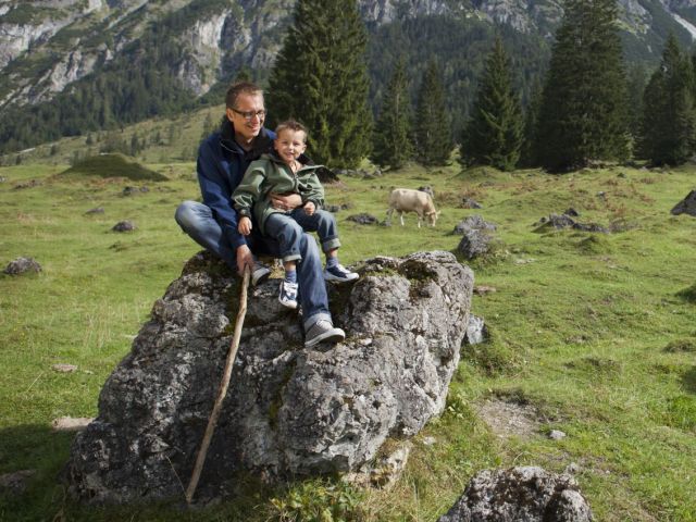 Vater sitzt mit Sohn auf großem Felsbrocken. Vater hält langen Stock in der Hand.