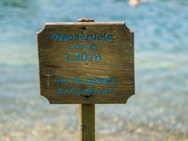 Nahaufname von Holzschild vor dem Badesee. Auf dem Schild steht: Wassertiefe mehr als 1,30m. Nur für geübte Schwimmer!