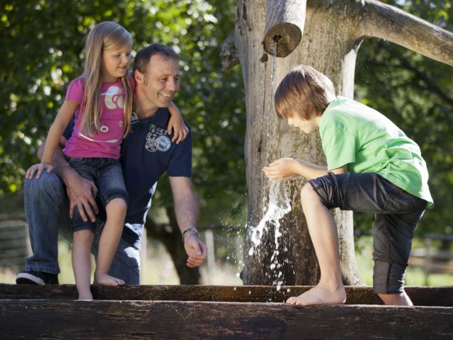 Vater, Tochter und Sohn stehen an einem Brunnen. Sohn trinkt Wasser und Vater hält Tochter im Arm.
