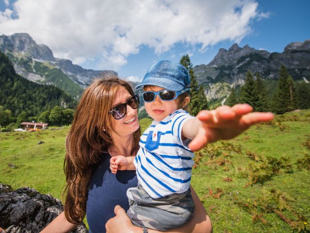 Sitting mother with sunglasses has little son with sunglasses on his arm. In the background is a mountain landscape.